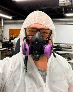 Asbestos worker wearing full PPE, including a respirator mask, protective suit, and safety glasses, in an industrial training environment.